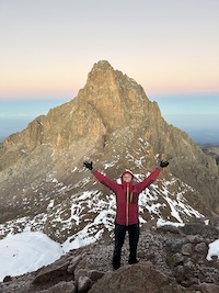 Hannah at the top of a mountain summit, arms raised victoriously.